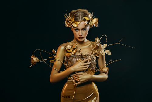 Woman wearing a gold dress and gold flower wreath holding gold flowers and staring intensely