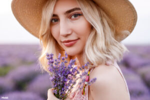 Woman with blonde hair wearing a straw hat smiling and holding a handful of purple flowers
