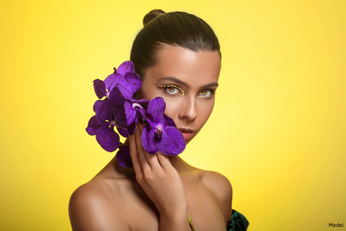 Brunette woman with hair pulled back in a bun holding a purple flower to her face in front of a yellow background