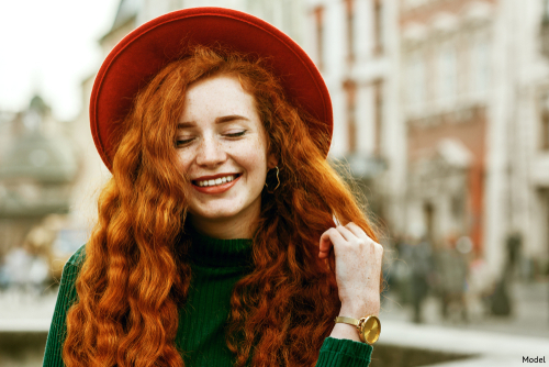 Smiling young woman with long, curly red hair wearing a green sweater and red hat outdoors