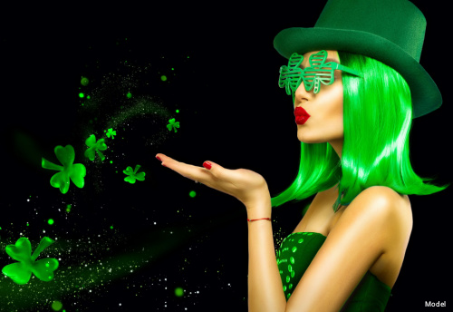 Woman in a green wig, green hat and shamrock shutter shades blowing shamrocks into the air